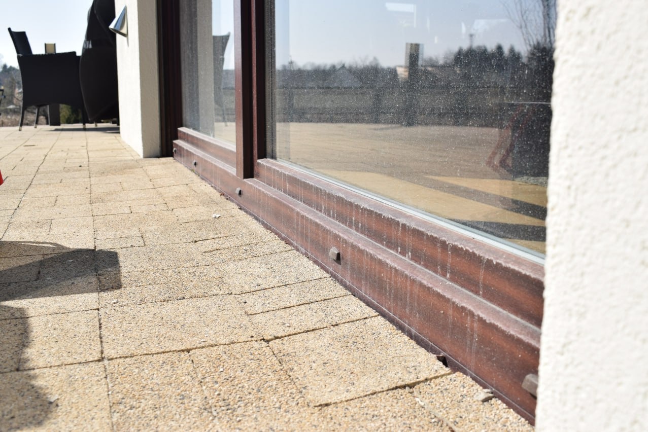 Brudne okno przesuwne z brązową ramą, widoczne z bliska od strony tarasu wyłożonego kostką, ślady po deszczu na szybie i ramie.