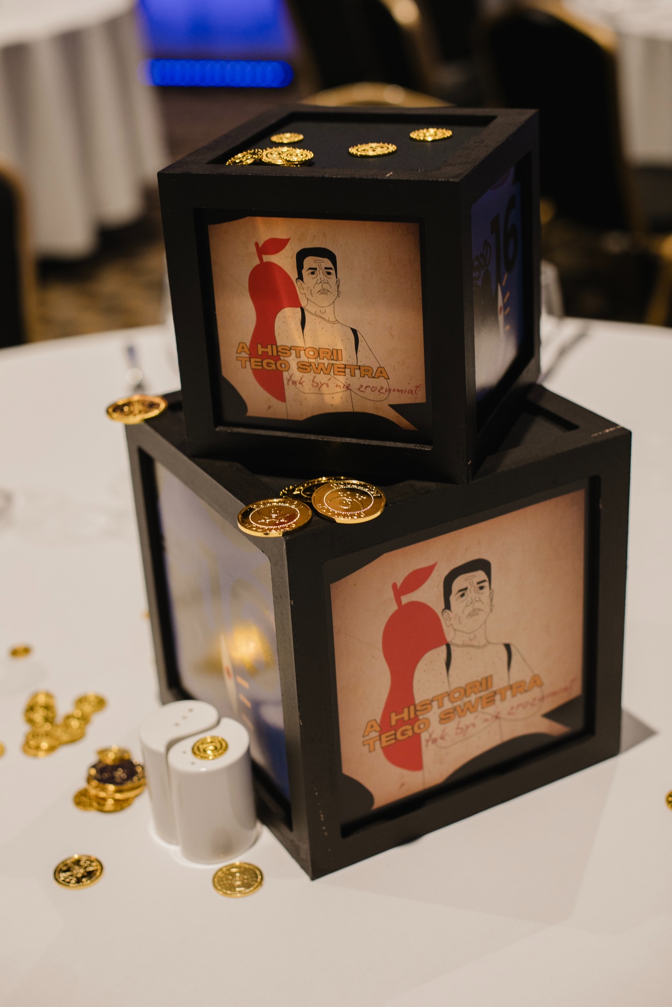 Dekoracja stołu na imprezie integracyjnej: dwa czarne, kwadratowe pudełka z grafikami i złotymi monetami, solniczka Yin-Yang, tworząca unikalny element wystroju.