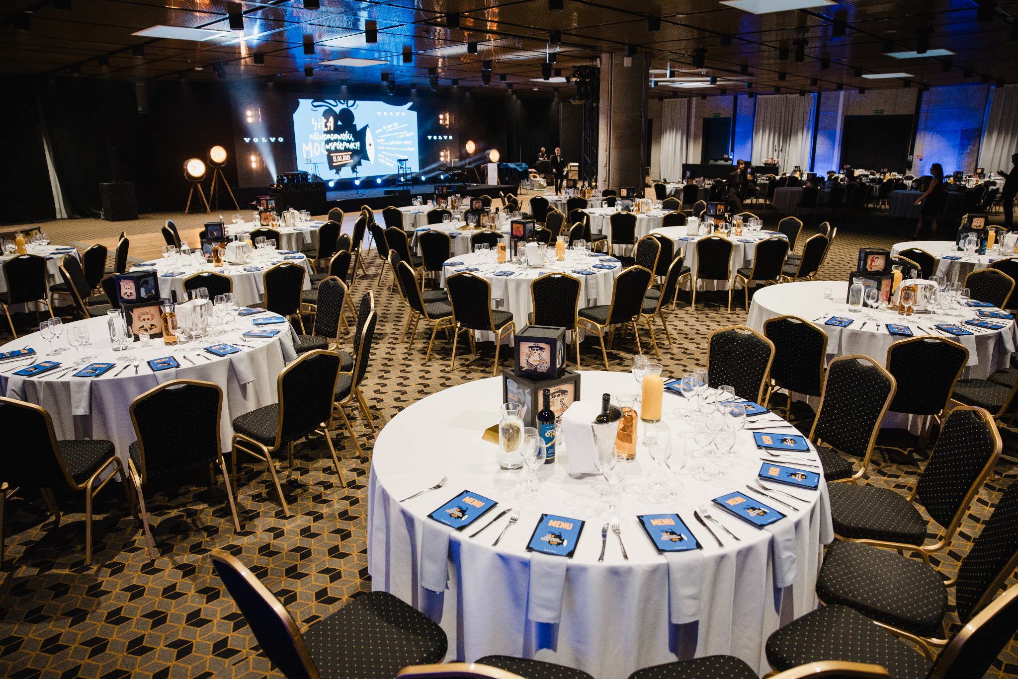 Sala bankietowa z okrągłymi stołami nakrytymi białymi obrusami i czarnymi krzesłami. Na scenie oświetlenie i ekran z logo. Przygotowania do imprezy.