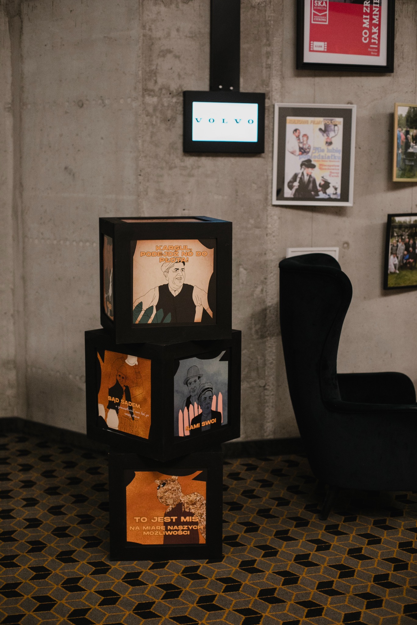 Dekoracja z czarnych sześcianów z grafikami filmowymi, obok czarny fotel, na ścianie plakaty filmowe i logo Volvo. Element wystroju wnętrza, prawdopodobnie na imprezie.