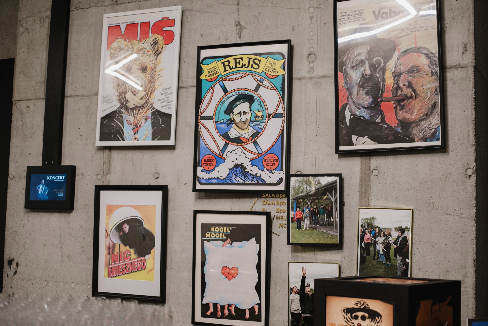 Ściana z betonu z kolekcją oprawionych plakatów filmowych, w tym 'Miś', 'Rejs', 'Vabank' i 'Kogel Mogel', tworzących eklektyczną galerię. Dodatkowo mały ekran z napisem 'Koncert'.