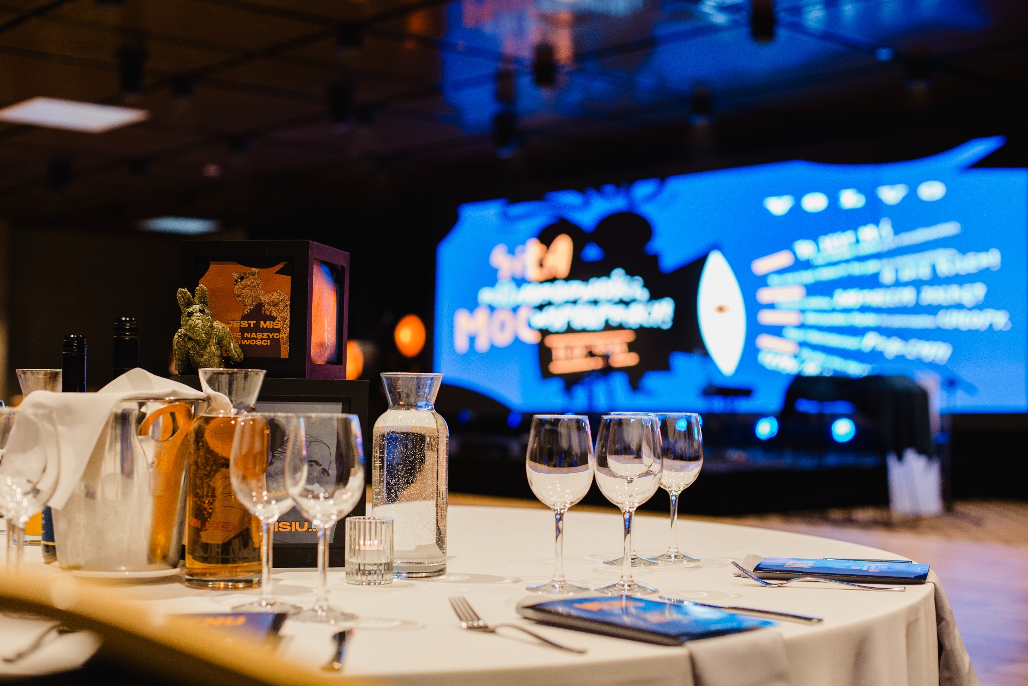 Uroczysty stół z kieliszkami, karafką i nagrodą na tle ekranu z logo Volvo, przygotowany na imprezę firmową. Dekoracje i oświetlenie tworzą elegancką atmosferę.