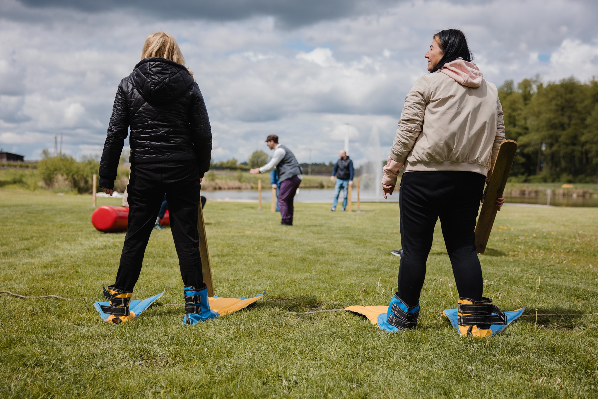 Uczestnicy w dziwnych butach rywalizują w nietypowej grze zespołowej na trawiastym terenie podczas imprezy integracyjnej na świeżym powietrzu w słoneczny dzień.