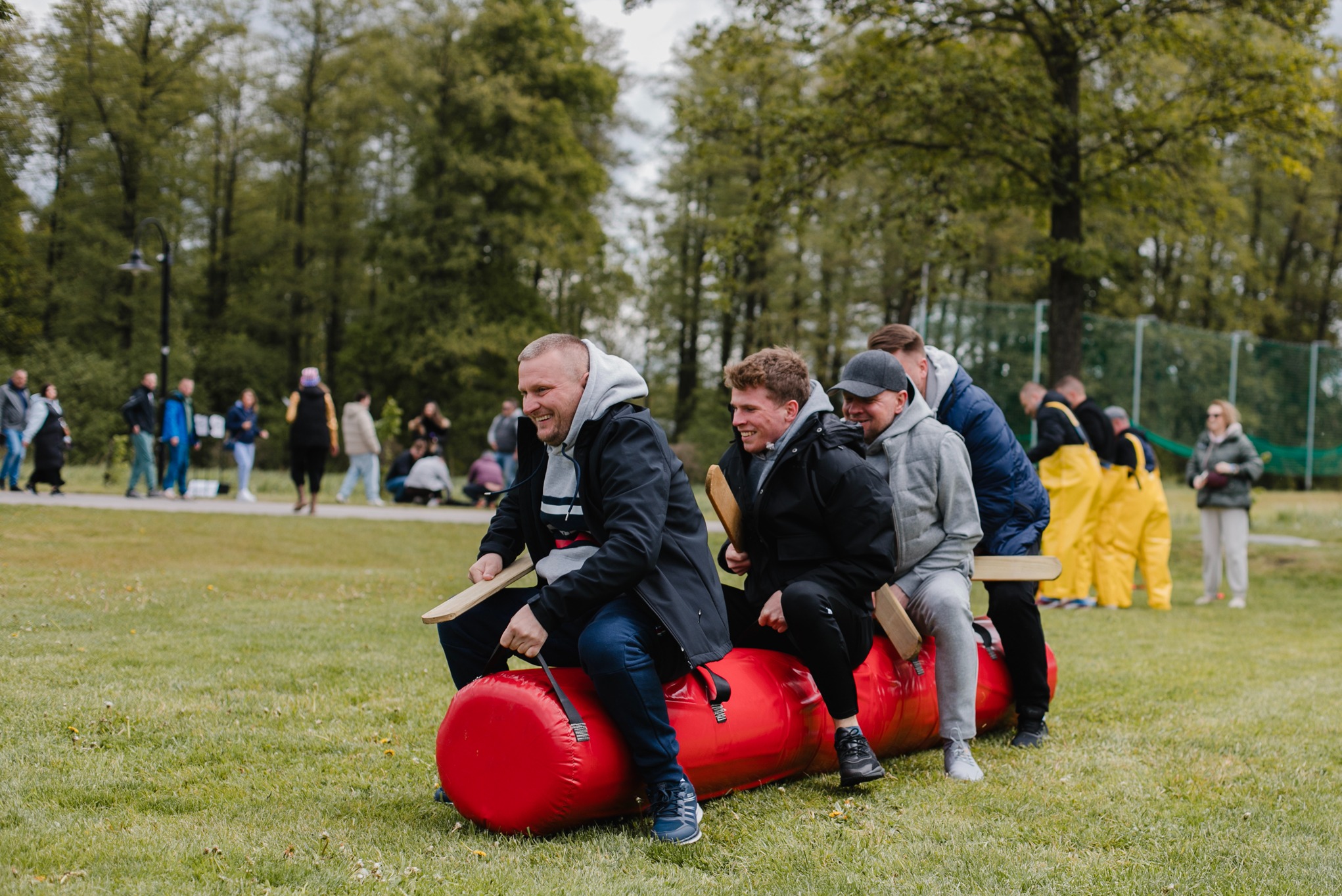 Grupa osób uśmiechnięta, siedzi na czerwonym walcu podczas konkurencji na trawie. W tle widoczni obserwatorzy i drzewa, co sugeruje imprezę integracyjną.