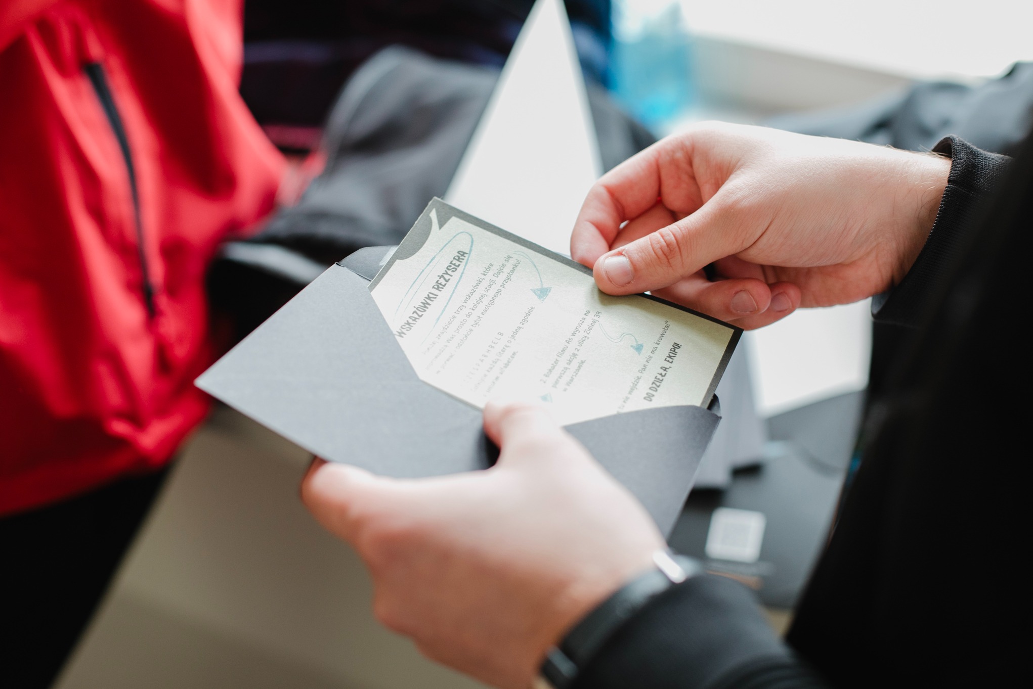 Otwarta, grafitowa koperta z kartką w dłoniach. Kartka z instrukcjami/wskazówkami do gry miejskiej lub imprezy integracyjnej. Widoczny fragment tekstu.