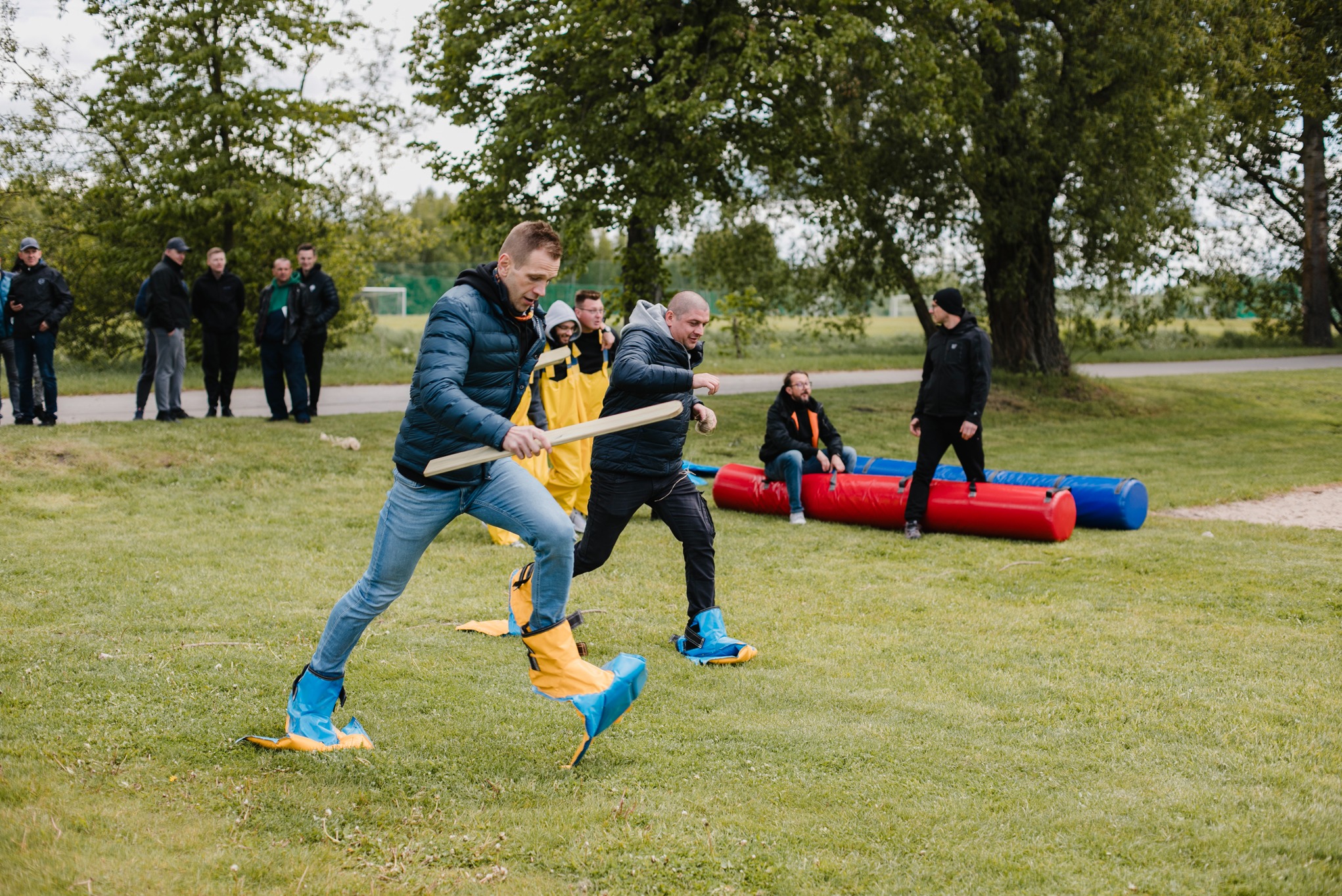 Uczestnicy biegną w zabawnych, kolorowych butach podczas imprezy integracyjnej na trawie. W tle grupa osób i dmuchane przeszkody.