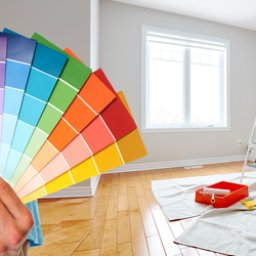 GLOBAL KOMPLEKS SPÓŁKA Z OGRANICZONĄ ODPOWIEDZIALNOŚCIĄ - Paleta kolorów trzymana w dłoni na tle przygotowanego do malowania jasnego pokoju z drewnianą podłogą, drabina i kuweta malarska na podłodze.