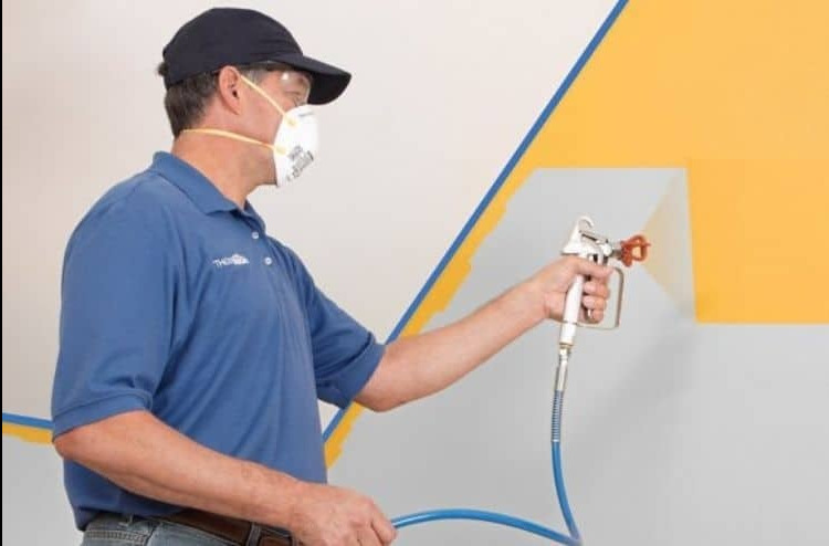 Pracownik w masce ochronnej aplikuje żółtą farbę natryskową na ścianę, zabezpieczoną niebieską taśmą malarską.