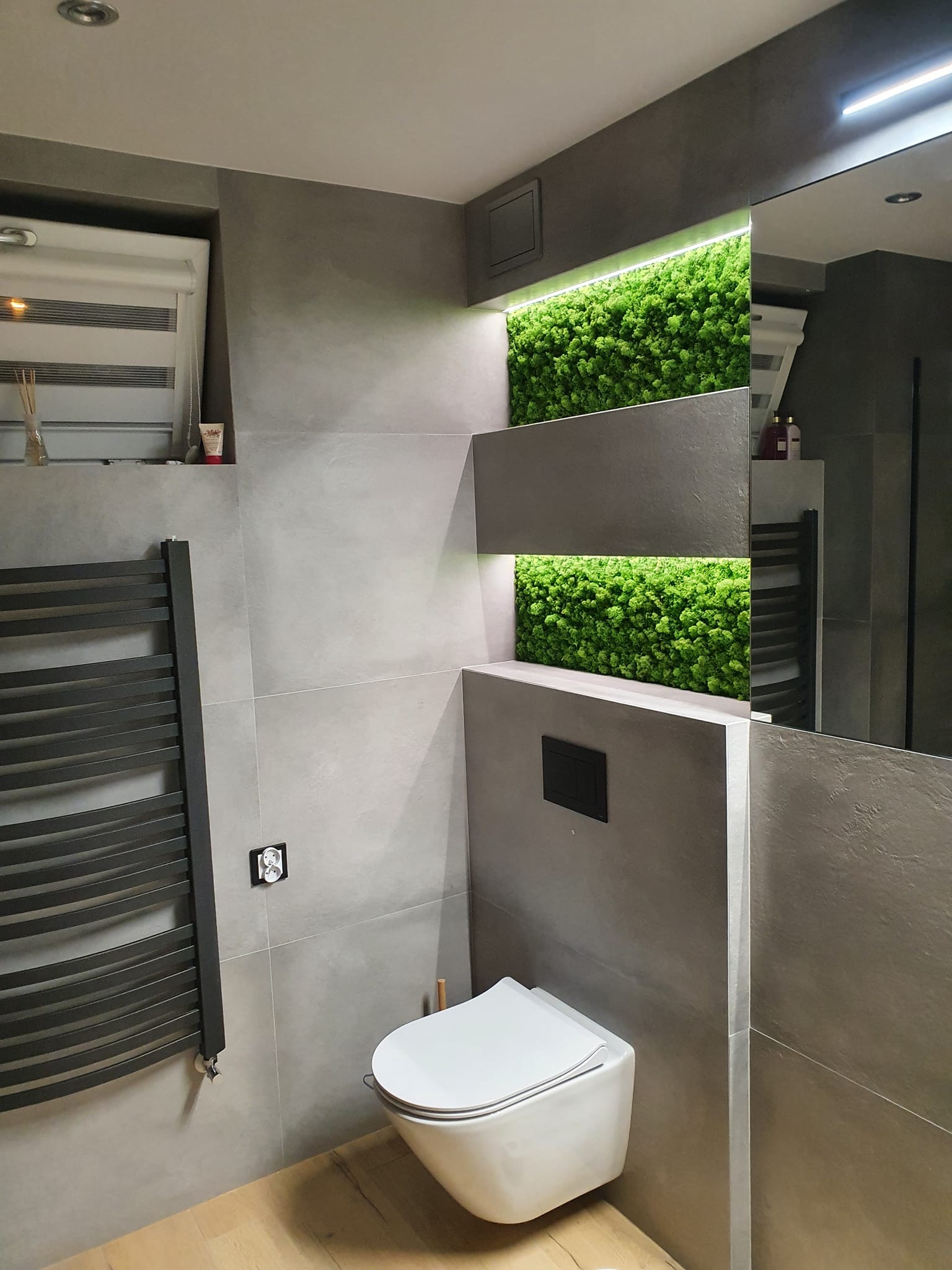 Nowoczesna łazienka z szarymi ścianami, podwieszaną toaletą, grzejnikiem drabinkowym i dekoracyjnym mchem podświetlonym LED w dwóch wnękach.