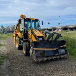 Żółta koparko-ładowarka JCB na żwirowej drodze, z widocznym wiaduktem w tle. Sprzęt budowlany gotowy do pracy.