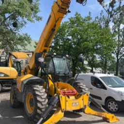 Żółty podnośnik teleskopowy JCB na placu budowy z wysuniętymi podporami, obok zaparkowany biały samochód dostawczy. W tle zieleń drzew i budynek.