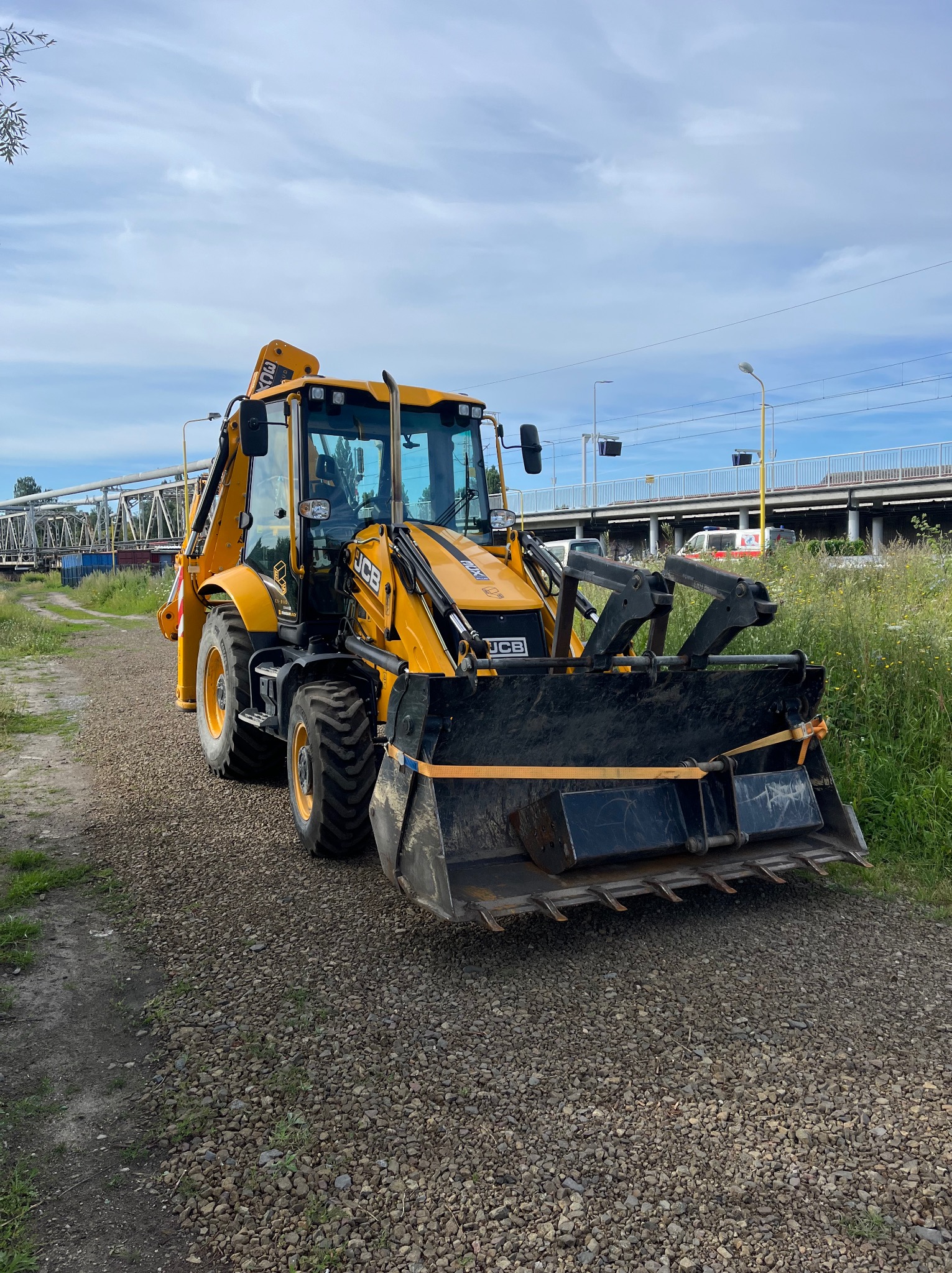 Żółta koparko-ładowarka JCB na żwirowej drodze, z widocznym wiaduktem w tle. Sprzęt budowlany gotowy do pracy.