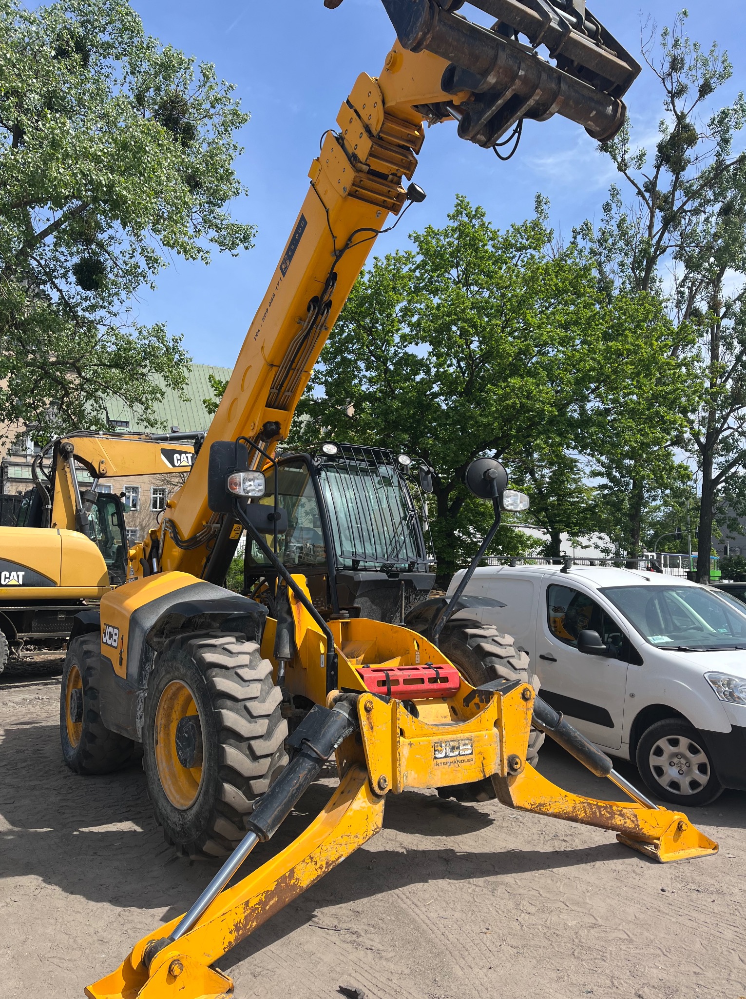 Żółty podnośnik teleskopowy JCB na placu budowy z wysuniętymi podporami, obok zaparkowany biały samochód dostawczy. W tle zieleń drzew i budynek.