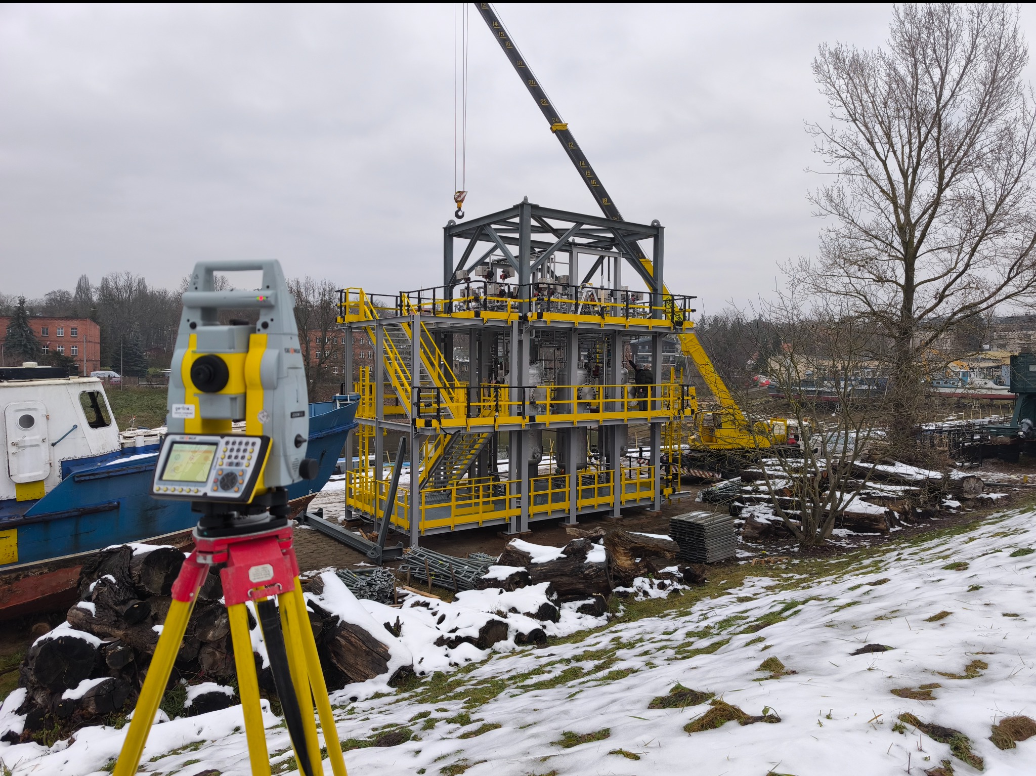 Pomiar geodezyjny konstrukcji stalowej z użyciem niwelatora na tle zimowego krajobrazu i dźwigu budowlanego. Prace pomiarowe w toku.