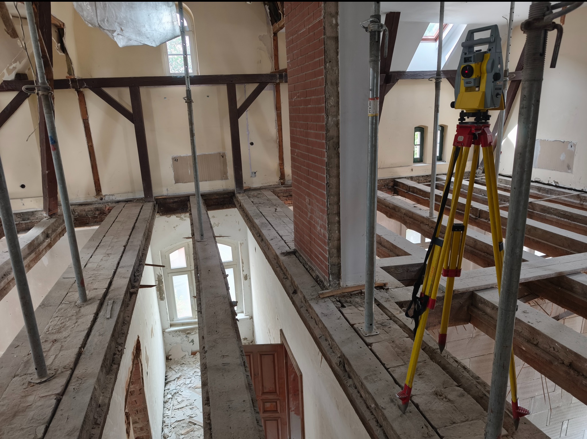 Żółty niwelator na statywie w surowym wnętrzu remontowanego poddasza z odsłoniętą konstrukcją drewnianą i ceglanym kominem. Widoczne podpory i okna.