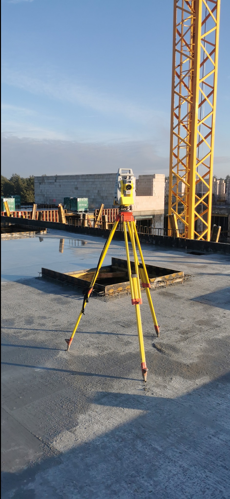 Żółty niwelator na trójnogu na betonowym stropie budowy, w tle dźwig i konstrukcja budynku. Pomiar geodezyjny na placu budowy.