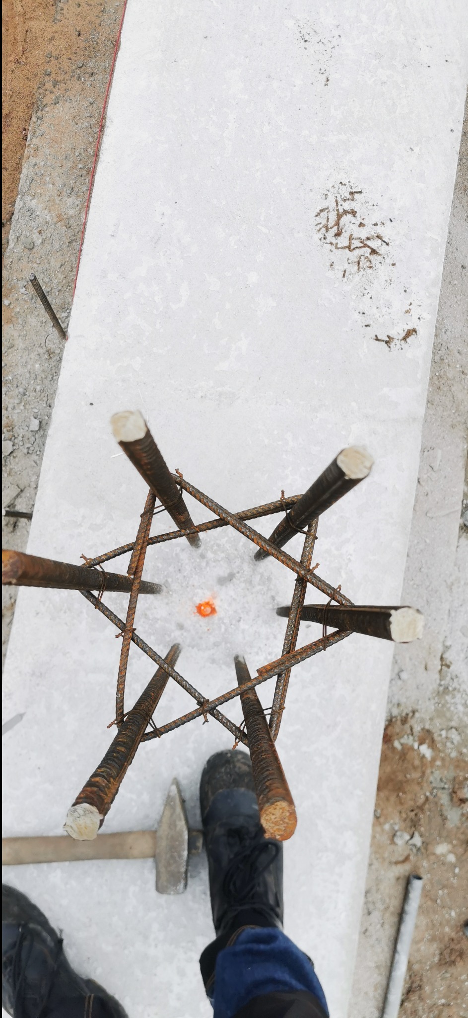 Zbrojenie słupa fundamentowego z zaznaczonym punktem centralnym, widok z góry. Obok leży młotek i widać fragment buta pracownika. Szalunek z betonu.