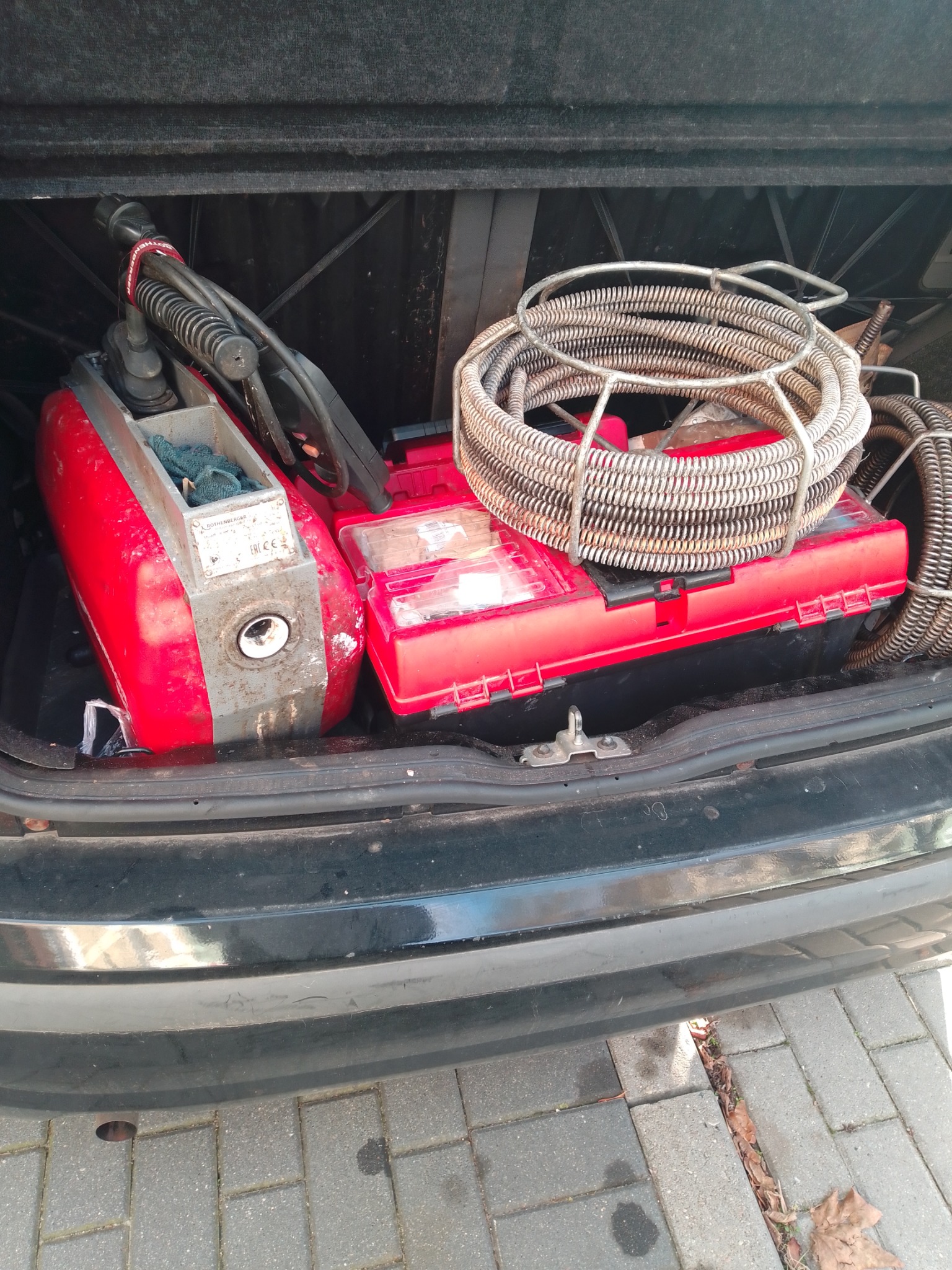 Sprzęt do udrażniania rur w bagażniku samochodu: czerwona maszyna Rothenberger, zwój sprężyny w metalowym koszu i czerwona skrzynka narzędziowa. Widok z góry, bagażnik otwarty.