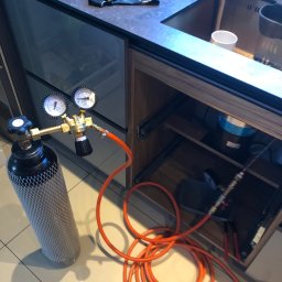 B&G SERVICE Beniamin Góźdź - Butla z gazem i reduktorem ciśnienia pod zlewem, pomarańczowy wąż prowadzi do instalacji w szafce.