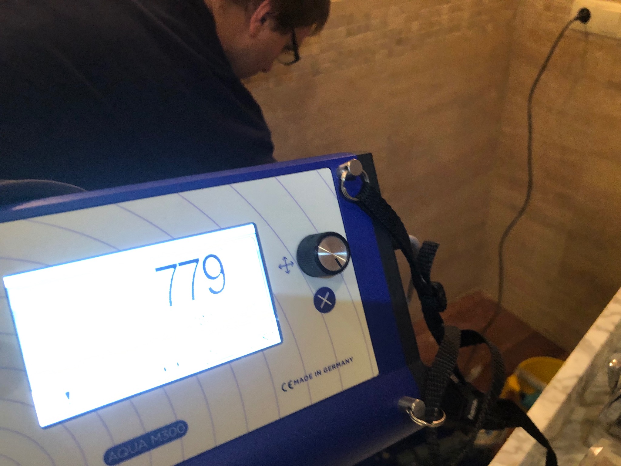Hydraulik w trakcie pomiaru parametrów wody za pomocą urządzenia Aqua M300 z wyświetlaczem wskazującym wartość 779, widoczna wtyczka w gniazdku.