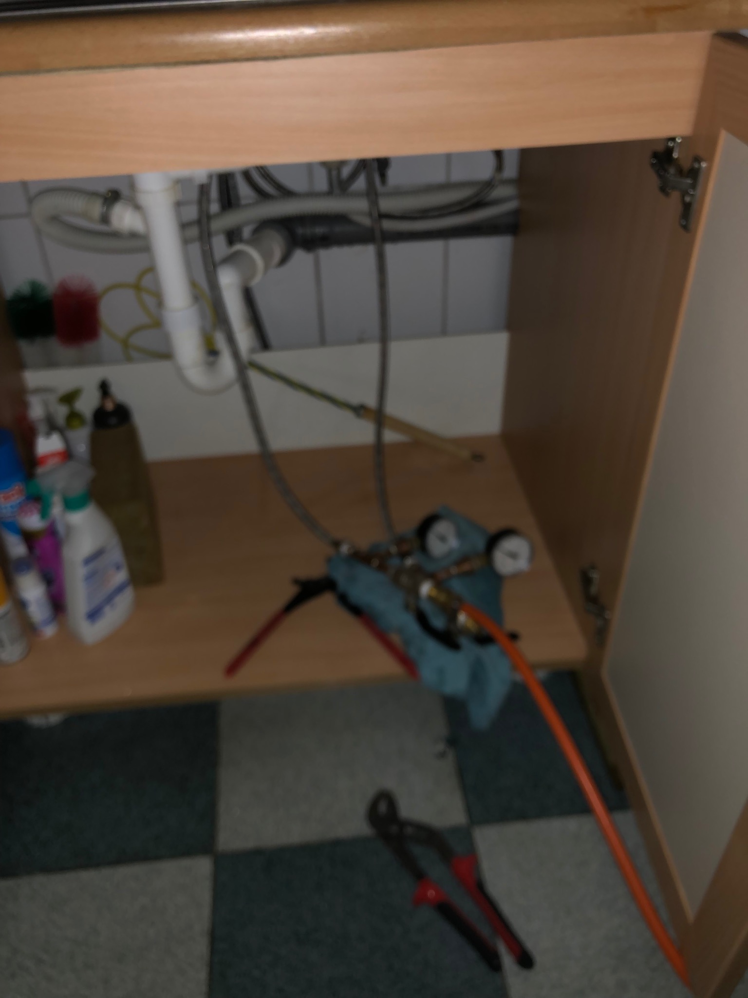 Podłączenie instalacji wodnej pod zlewem w szafce, widoczne rury, węże, manometry oraz narzędzia hydrauliczne na podłodze z płytek.