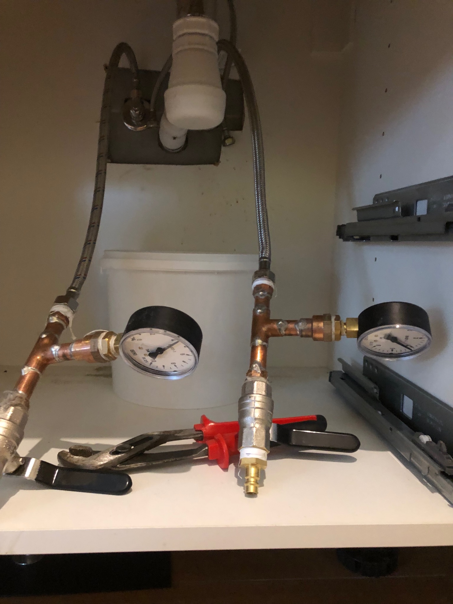 Miedziane rury z manometrami pod zlewem, widoczne narzędzia hydrauliczne i biały pojemnik w tle.