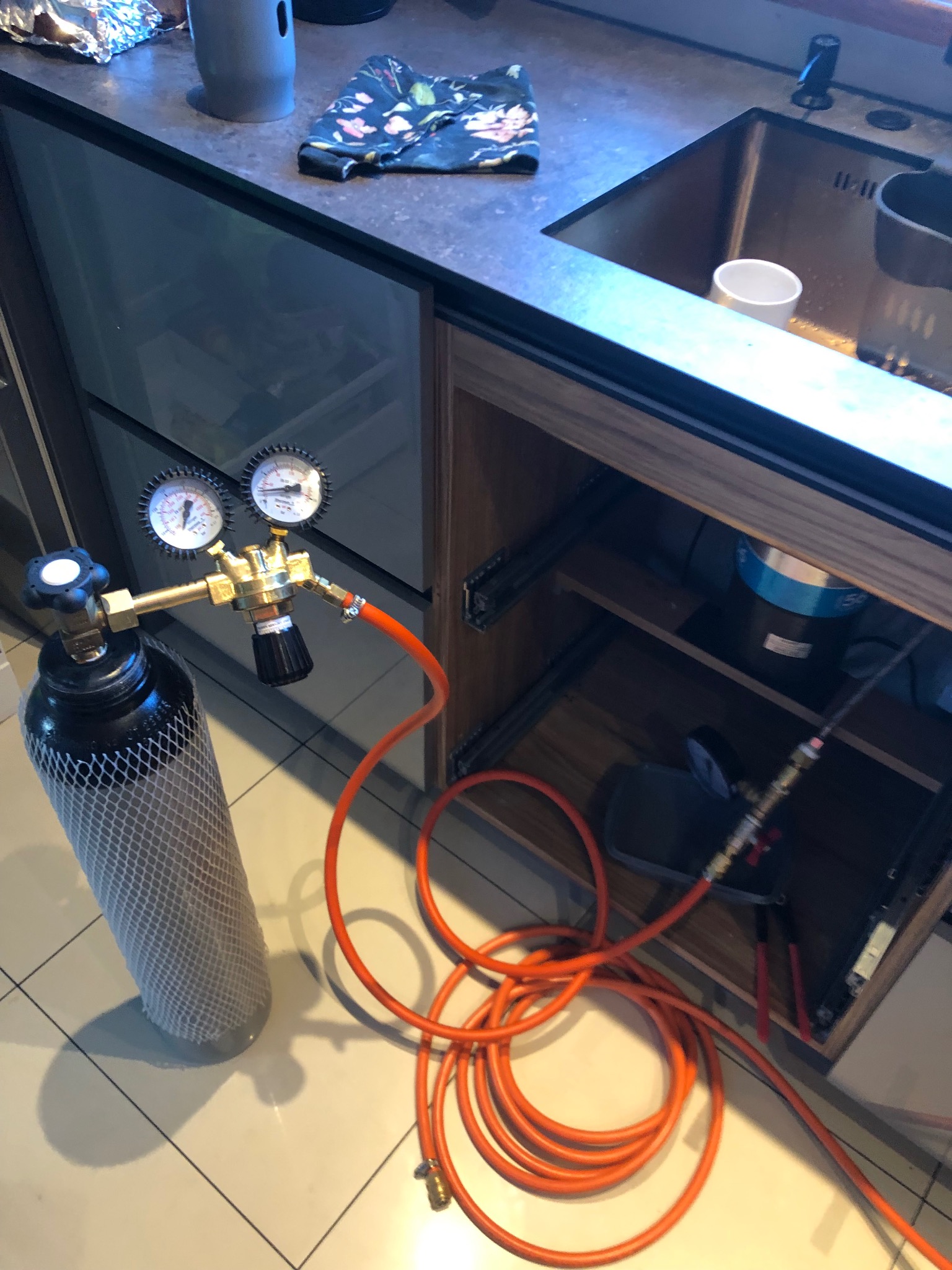 Butla z gazem i reduktorem ciśnienia pod zlewem, pomarańczowy wąż prowadzi do instalacji w szafce.