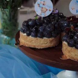 Tartaletki z borówkami na drewnianej paterze, ozdobione topperami z misiem i balonami, na tle błękitnego tiulu i kwiatów w wazonie.