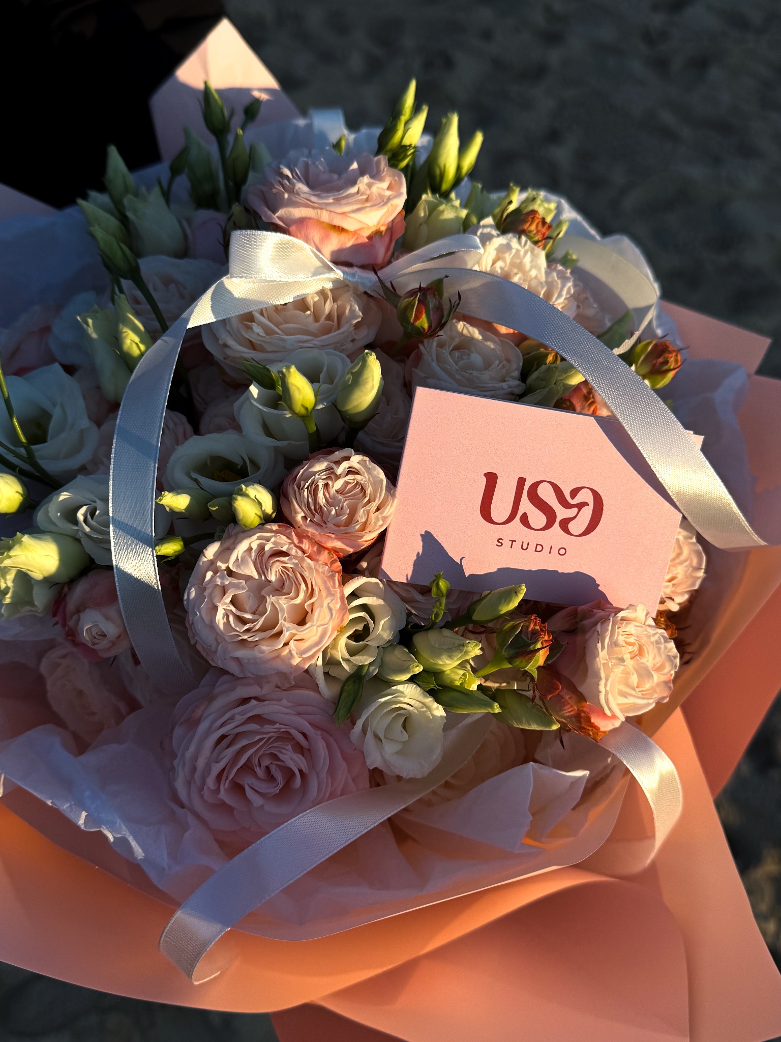 Delikatny bukiet z różowych i białych róż oraz eustomy, przewiązany satynową wstążką, z dołączonym wizytownikiem 'US9 Studio' w pastelowym, różowym opakowaniu.