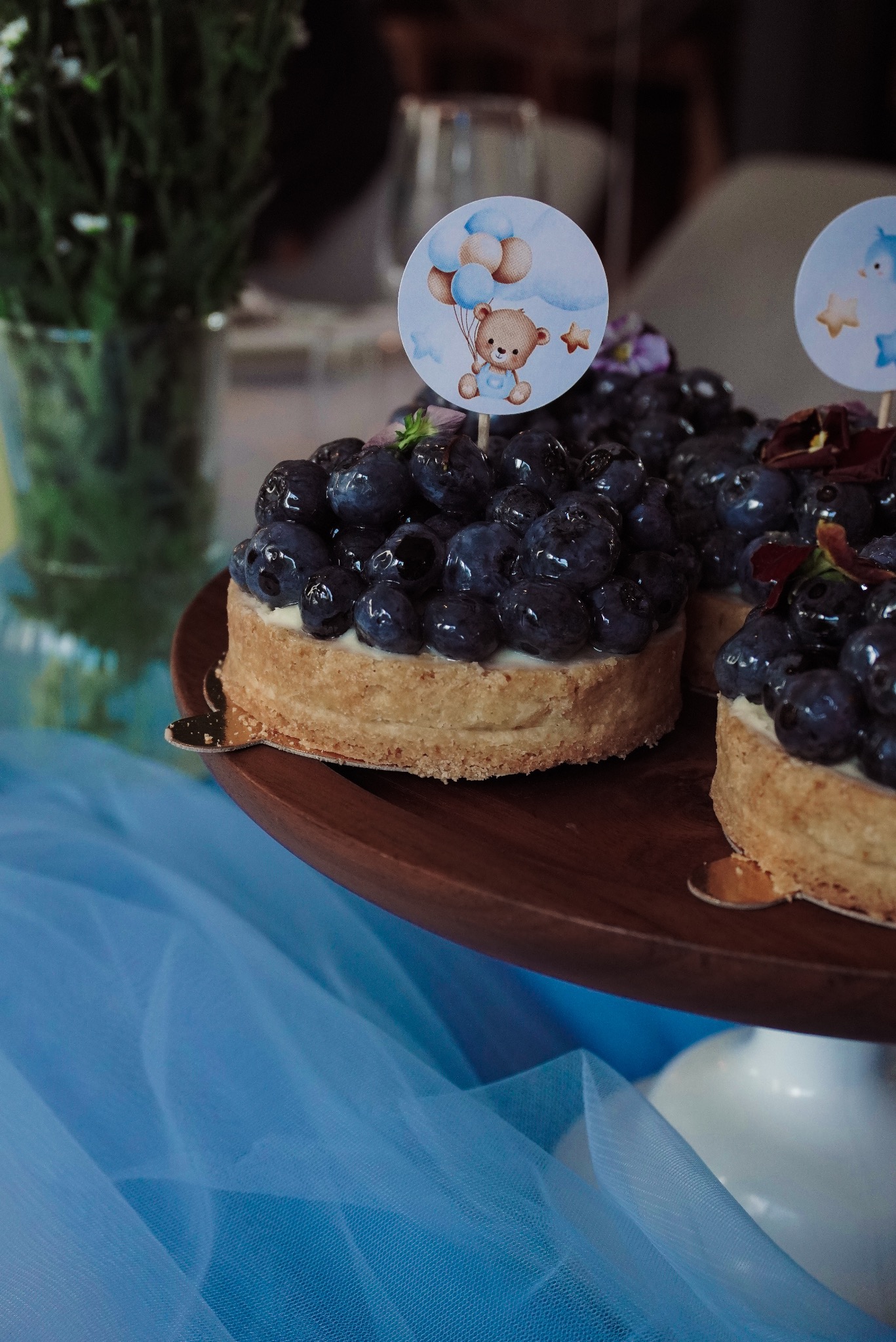Tartaletki z borówkami na drewnianej paterze, ozdobione topperami z misiem i balonami, na tle błękitnego tiulu i kwiatów w wazonie.