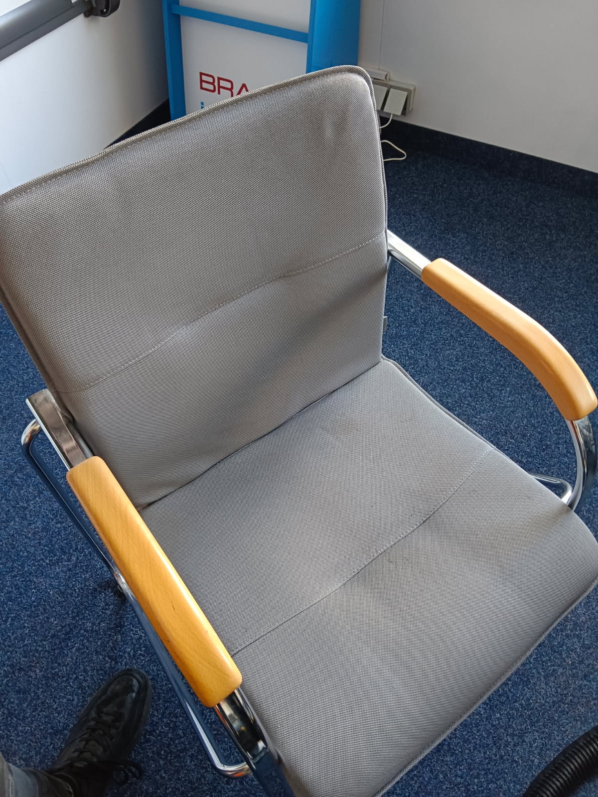 Szary fotel biurowy z drewnianymi podłokietnikami na tle niebieskiej wykładziny. Widoczne ślady użytkowania na siedzisku, sugerujące potrzebę czyszczenia tapicerki.
