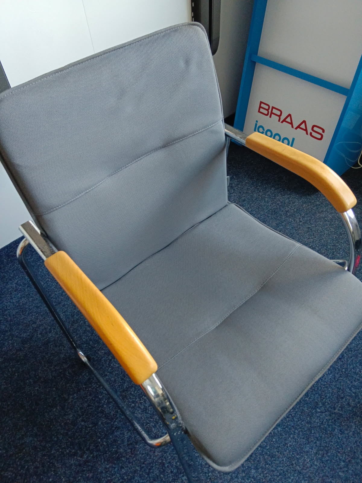 Szary fotel biurowy z drewnianymi podłokietnikami i chromowaną ramą, ustawiony na niebieskiej wykładzinie. Widoczny fragment reklamy Braas w tle.