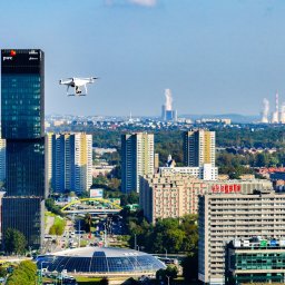 Let It Drone - Dron z kamerą w locie nad panoramą miasta z wieżowcami, kominami elektrowni w oddali i charakterystyczną halą widowiskowo-sportową w pierwszym planie.