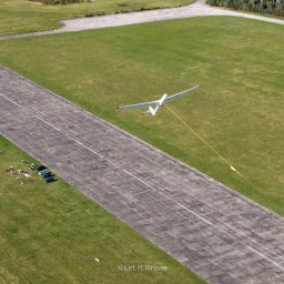 Let It Drone - Widok z góry na szybowiec holowany za pomocą liny nad pasem startowym, z grupą osób i samochodami na trawiastym poboczu.