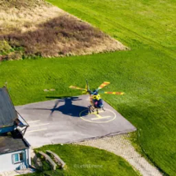 Żółto-czerwony helikopter ląduje na betonowym lądowisku z żółtym okręgiem, widok z góry na zielone pola i budynek z szarymi ścianami i ciemnym dachem.