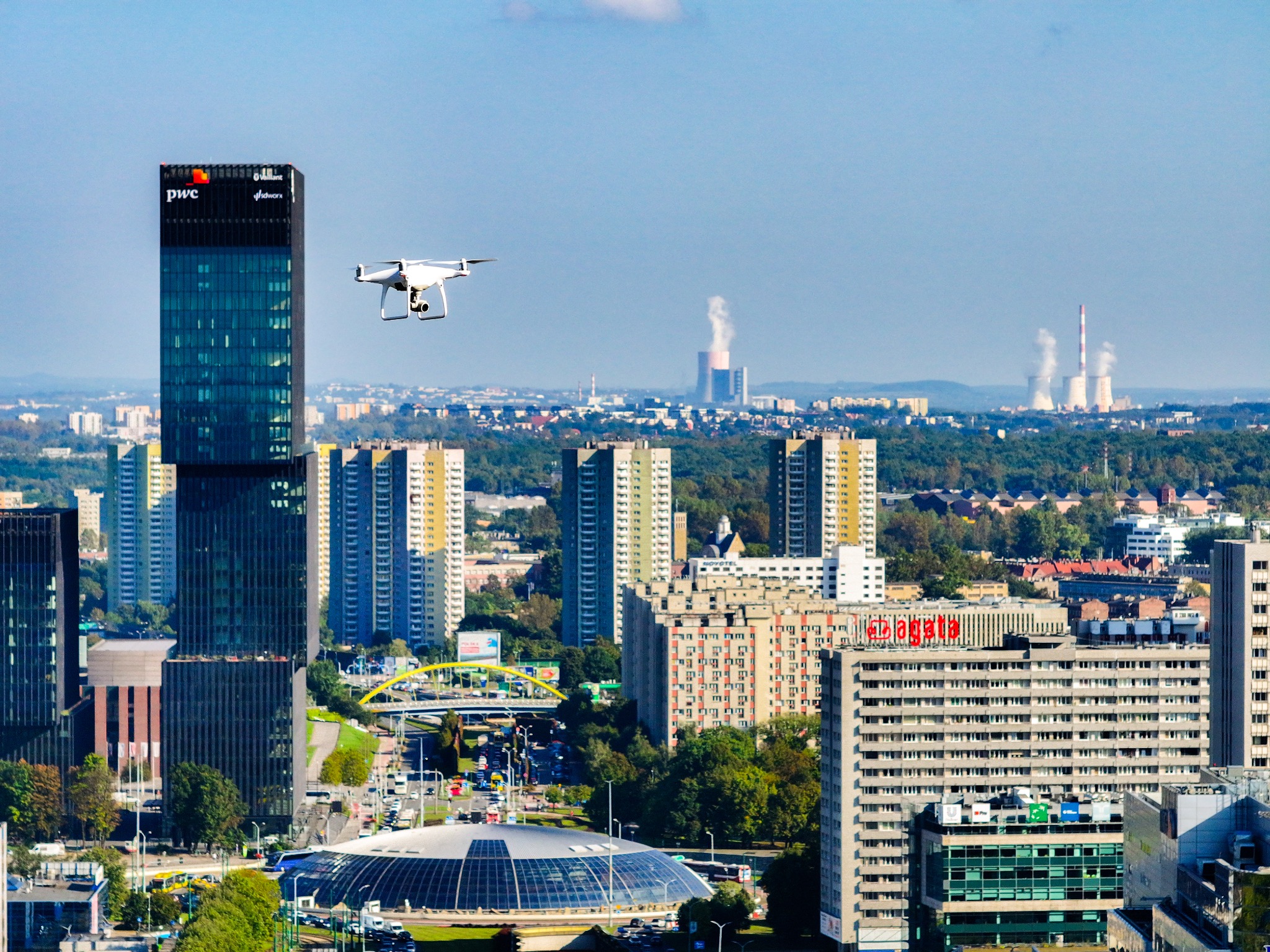 Dron z kamerą w locie nad panoramą miasta z wieżowcami, kominami elektrowni w oddali i charakterystyczną halą widowiskowo-sportową w pierwszym planie.