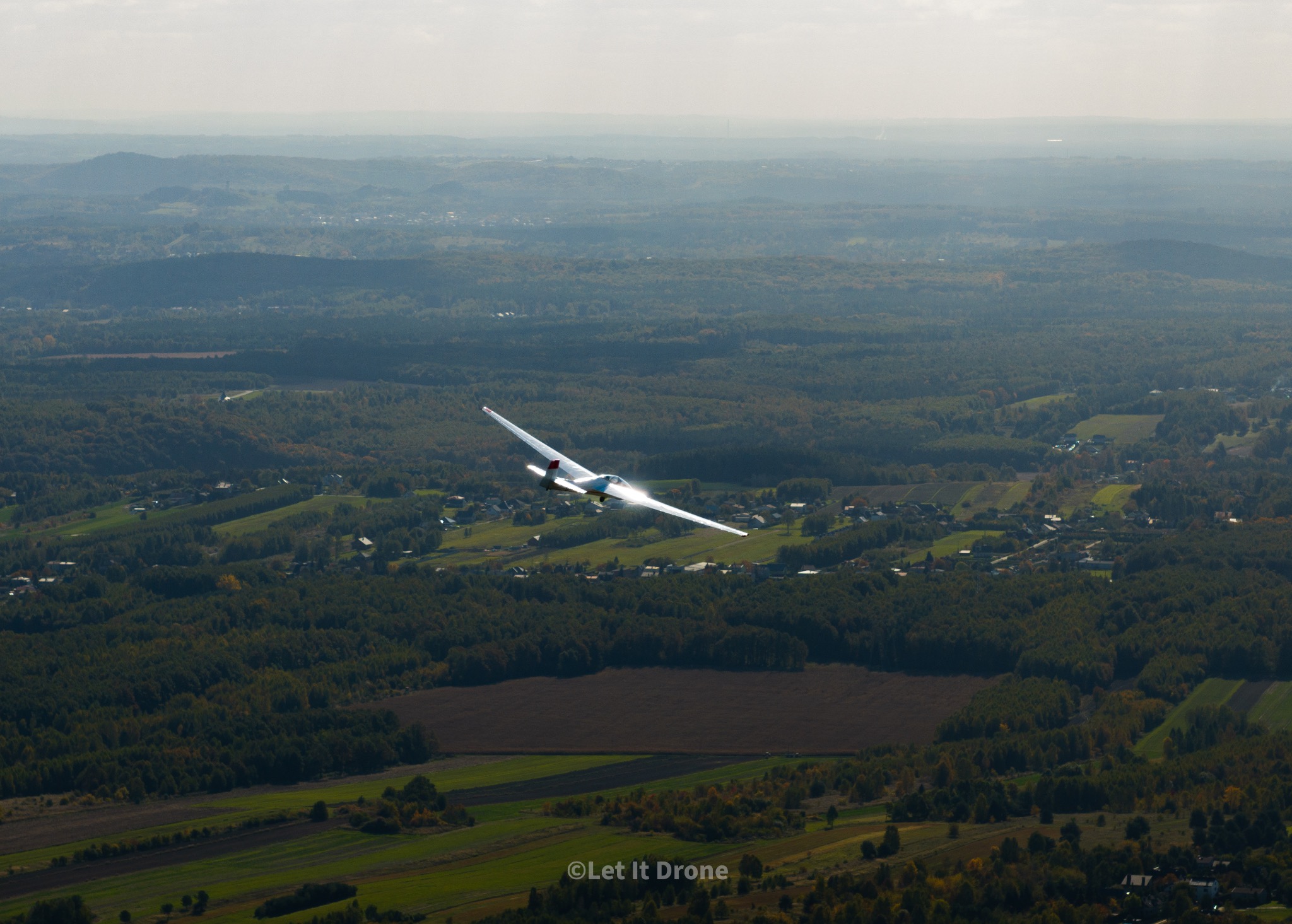 Szybowiec w locie nad pagórkowatym, zalesionym terenem, widok z lotu ptaka, lekka mgła w oddali.