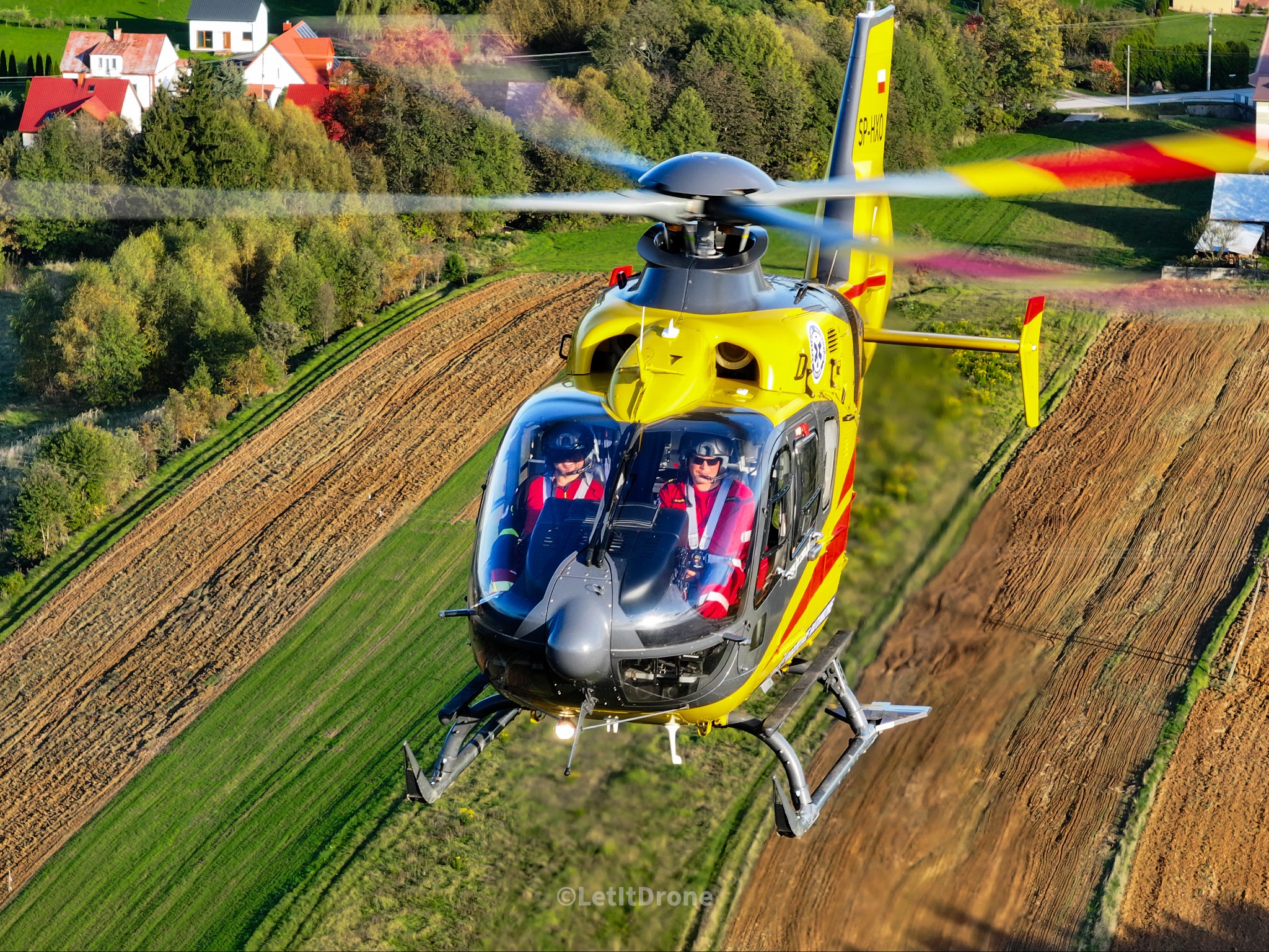 Żółto-czarny helikopter w locie nad polami uprawnymi i zabudowaniami, widoczni dwaj piloci w kabinie, śmigła w ruchu, ujęcie z przodu.