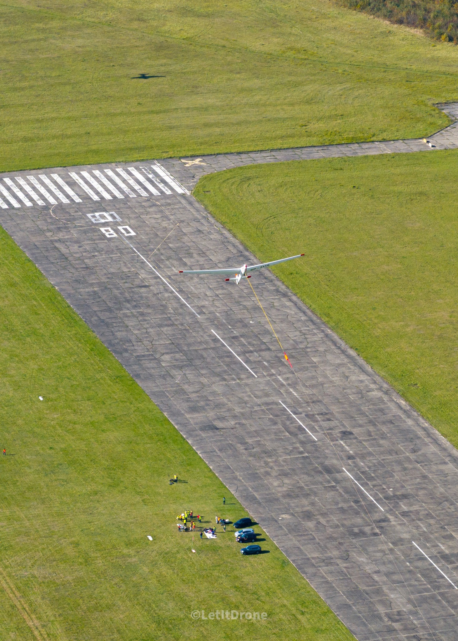 Szybowiec holowany na pasie startowym, widok z drona, grupa osób obserwująca na trawie obok zaparkowanych samochodów.