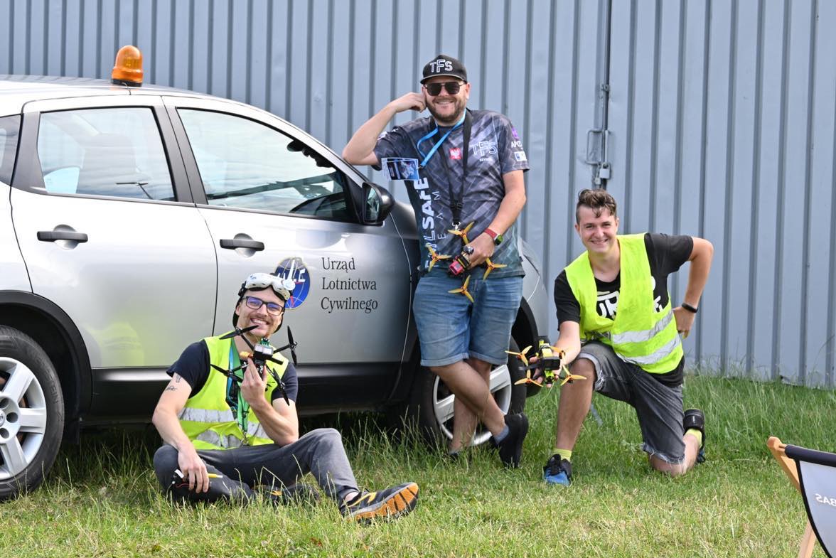 Trzech mężczyzn w kamizelkach odblaskowych pozuje z dronami wyścigowymi na tle samochodu Urzędu Lotnictwa Cywilnego i szarego blaszaka.