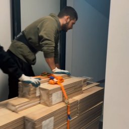 Serhii Kovela Przewóz - Pracownik w uprzęży transportuje wózkiem palety paneli podłogowych przez próg drzwi. Widoczne rękawice robocze i pomarańczowe pasy zabezpieczające ładunek.