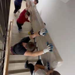 Serhii Kovela Przewóz - Czterech mężczyzn w rękawicach wnosi po schodach duży, owinięty mebel. Widok z góry, klatka schodowa z jasnymi ścianami i metalową barierką.