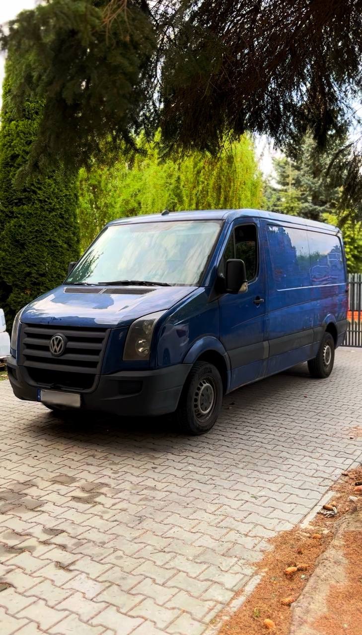 Niebieski van Volkswagen Crafter, zaparkowany na brukowanej nawierzchni, z zielonymi krzewami w tle. Widok z przodu i boku pojazdu, zarys ogrodzenia w tle.