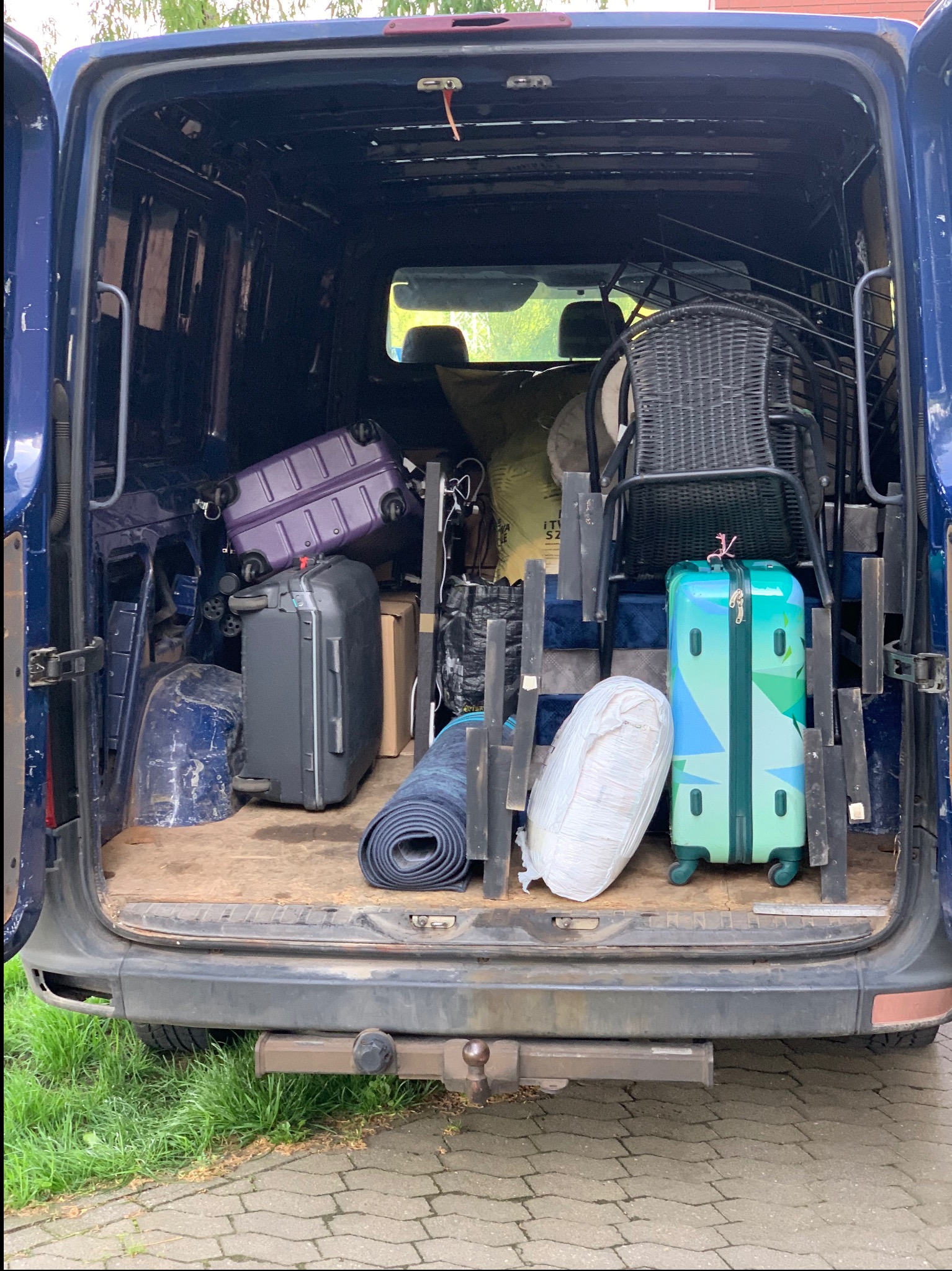 Załadunek busa: walizki (fioletowa, szara, turkusowa), zwinięty dywan, krzesło wiklinowe i inne przedmioty wewnątrz niebieskiego busa, widok z tyłu, trawa i kostka brukowa.