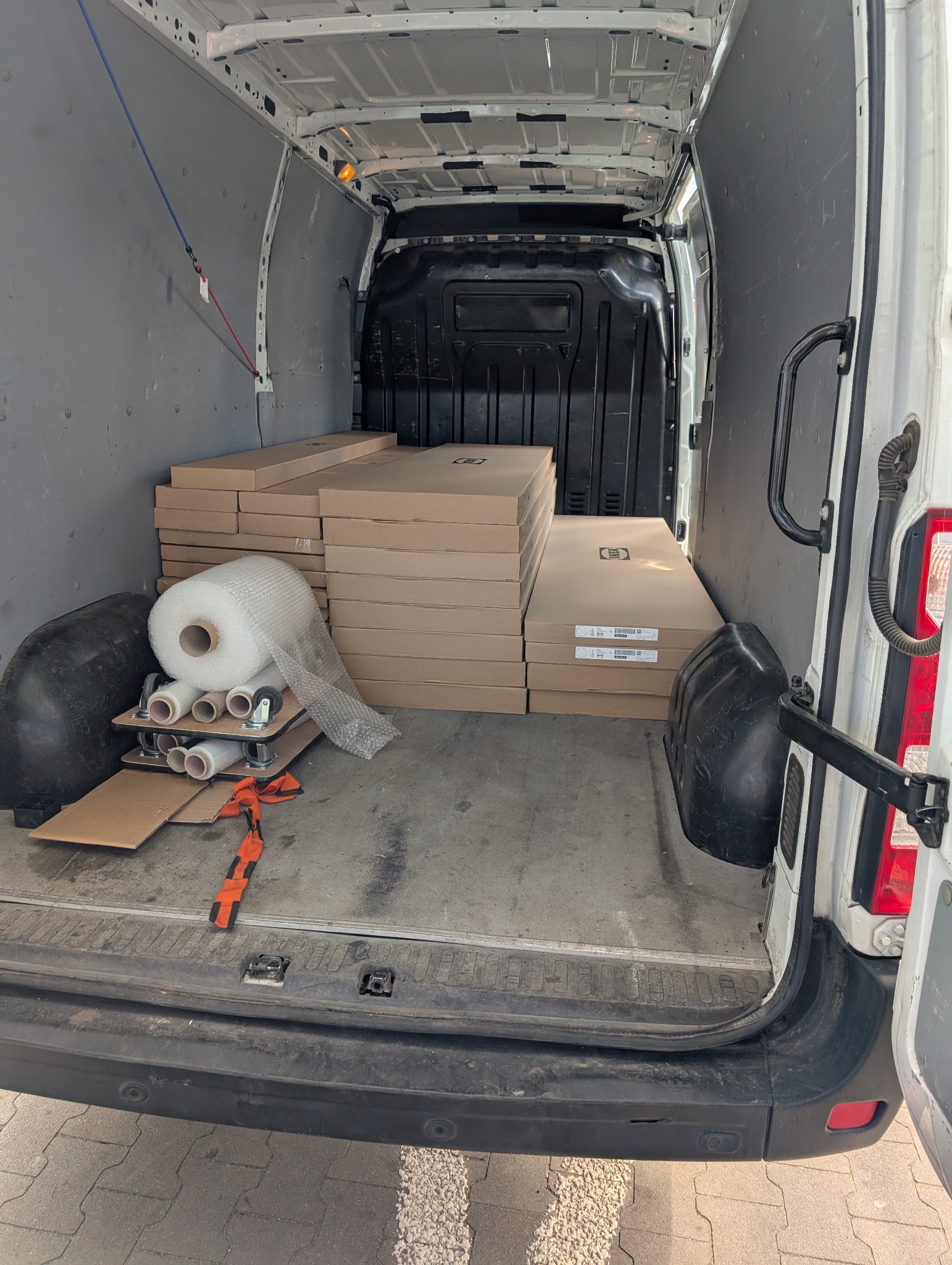 Wnętrze białego busa z załadunkiem: kartony, folia bąbelkowa i akcesoria do zabezpieczenia towaru. Widok od tyłu pojazdu, otwarte drzwi.