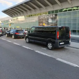 Czarny bus Opel Vivaro i auto osobowe na parkingu lotniska, z widocznym terminalem i reklamą DEFENDER. Usługi transportowe, transfer z lotniska.