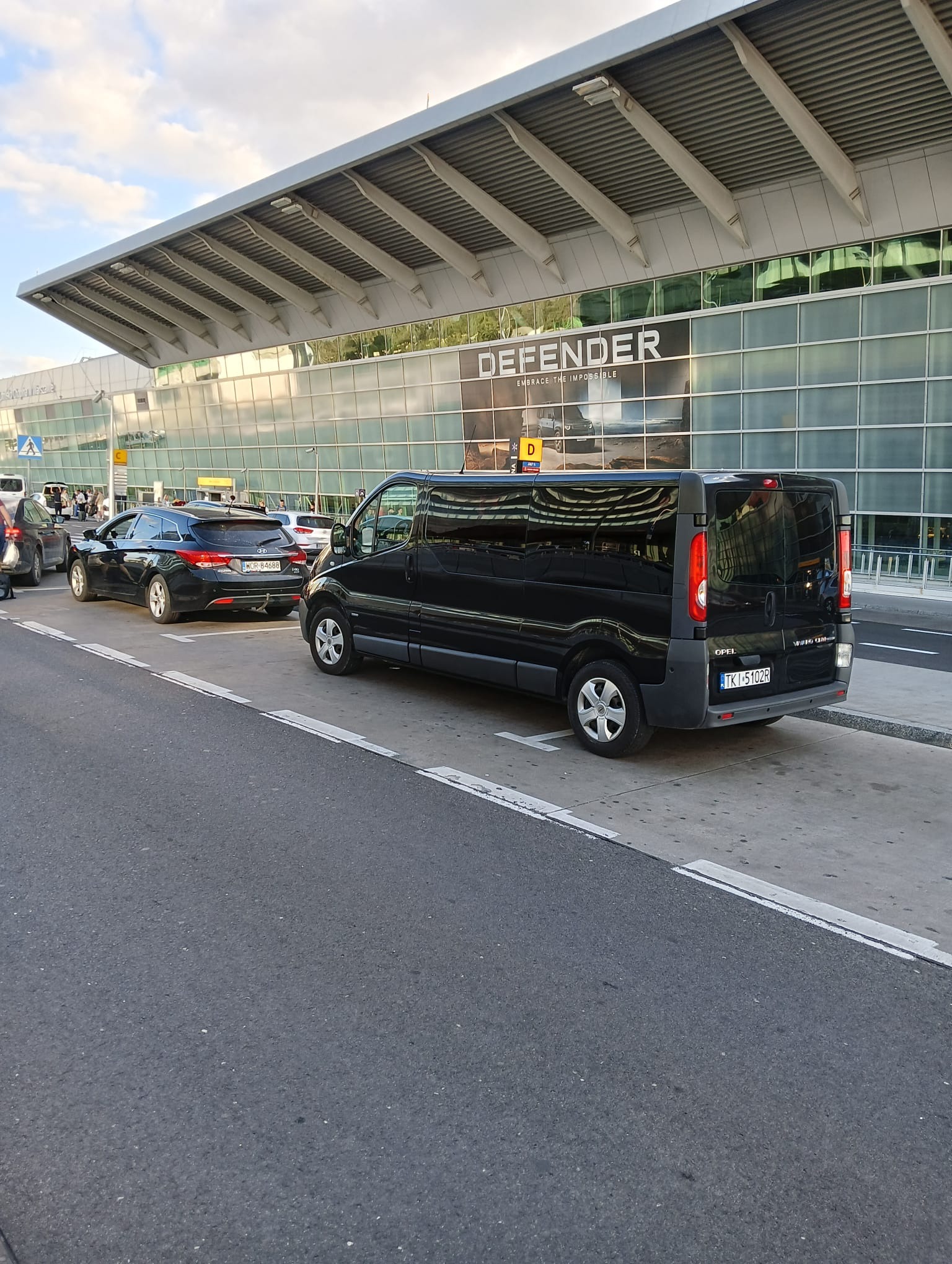 Czarny bus Opel Vivaro i auto osobowe na parkingu lotniska, z widocznym terminalem i reklamą DEFENDER. Usługi transportowe, transfer z lotniska.