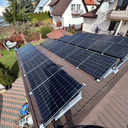 Profitenergy Spółka z ograniczoną odpowiedzialnością - Rząd paneli słonecznych na brązowym dachu domu jednorodzinnego, widok z góry, w tle zabudowa mieszkalna i zieleń.