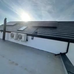 Panele słoneczne na dachu z dachówką, dwa klimatyzatory zamontowane na białej ścianie budynku, widoczne rury spustowe i okno dachowe, jasne słońce na niebie.