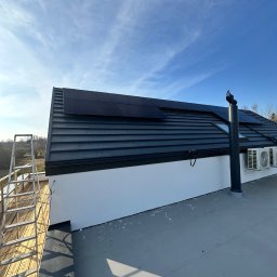 Profitenergy Spółka z ograniczoną odpowiedzialnością - Czarny dach z panelami fotowoltaicznymi i kominem wentylacyjnym w słoneczny dzień, widoczne dwa klimatyzatory na białej ścianie oraz drabina na drewnianym tarasie.